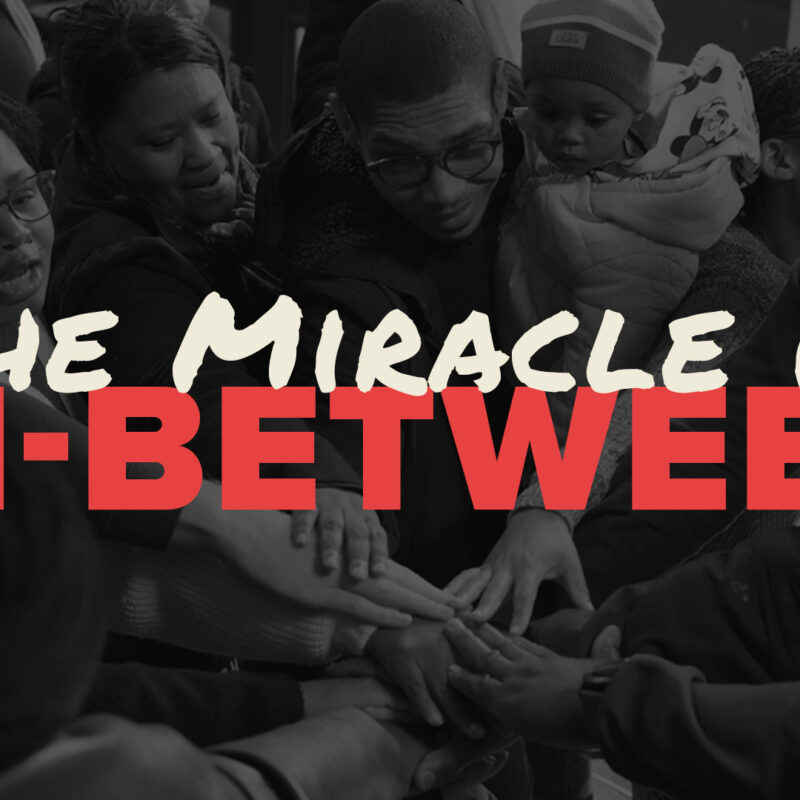 A picture of a group of people huddling together with the text "The Mieacle is In-Between" overlaid on it.