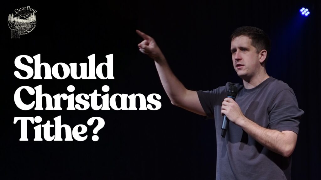 A man in a grey t-shirt stands on a dark stage, holding a microphone in his left hand and pointing upwards and to the right with his right hand. He has short, dark hair and is looking in the direction he is pointing. To the left of the image, the words "Should Christians Tithe?" are written in large white font. In the top left corner, there is a small circular logo with "Overflow" written above a stylized image of mountains and a river, matching the "Overflow" logo from the previous image. The background is dark, likely a stage or auditorium.