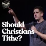 A man in a grey t-shirt stands on a dark stage, holding a microphone in his left hand and pointing upwards and to the right with his right hand. He has short, dark hair and is looking in the direction he is pointing. To the left of the image, the words "Should Christians Tithe?" are written in large white font. In the top left corner, there is a small circular logo with "Overflow" written above a stylized image of mountains and a river, matching the "Overflow" logo from the previous image. The background is dark, likely a stage or auditorium.