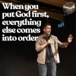 A man, Pastor Josh, is on a stage holding a microphone, actively speaking with one hand raised. He is wearing a light brown overshirt and dark jeans. Large white text on the left side of the image reads: "When you put God first, everything else comes into order". In the top right corner, there's a circular logo with the word "Overflow" and a mountain/tree design.