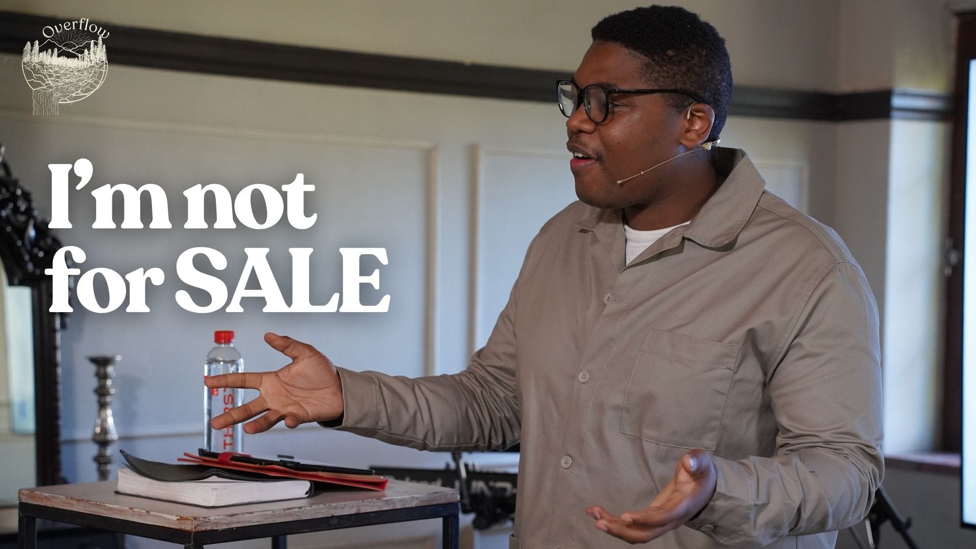 A young black man with glasses is speaking enthusiastically in a church setting. He is wearing a beige jacket and a microphone headset. He stands behind a small wooden table with a water bottle and a bible on it, gesturing with both hands. The text "I'm not for SALE" is prominently displayed on the left side of the image, with a logo for "Overflow" in the top left corner.