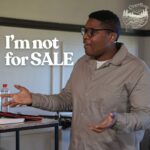 A young black man with glasses is speaking enthusiastically in a church setting. He is wearing a beige jacket and a microphone headset. He stands behind a small wooden table with a water bottle and a bible on it, gesturing with both hands. The text "I'm not for SALE" is prominently displayed on the left side of the image, with a logo for "Overflow" in the top right corner.