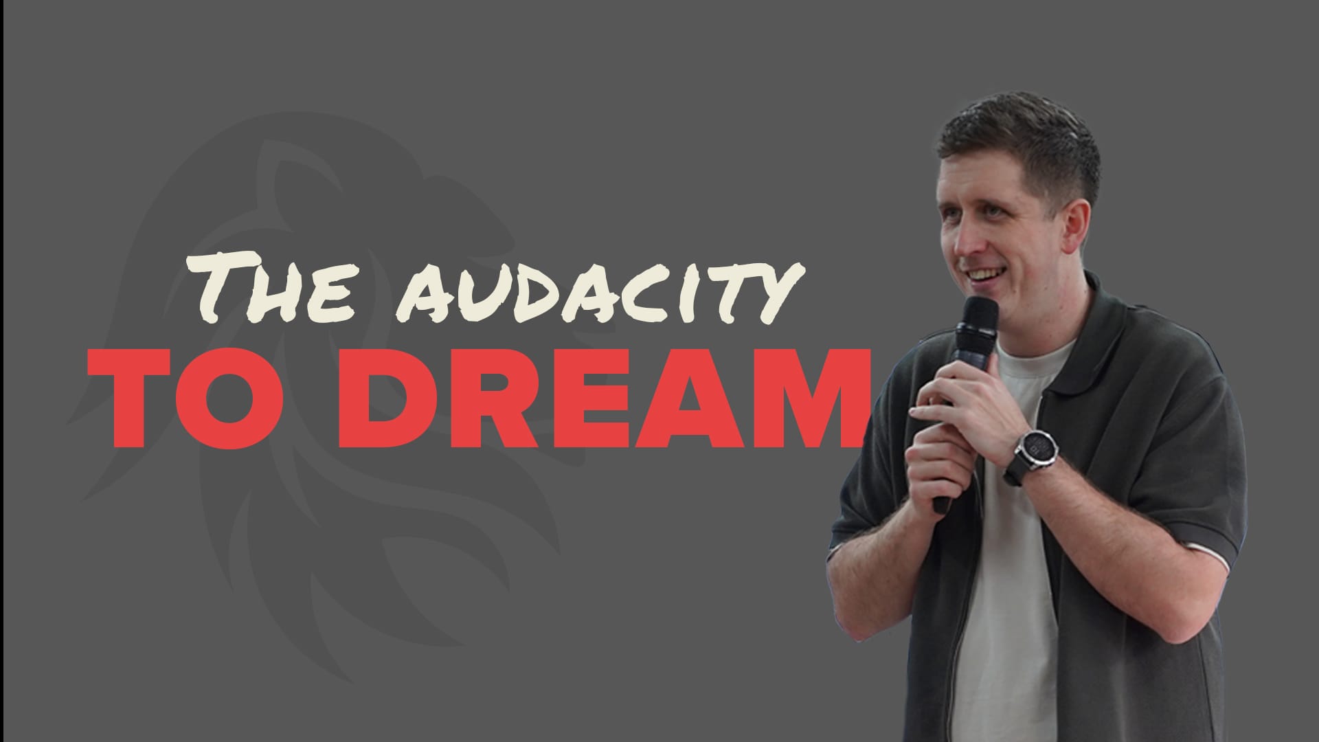 A horizontal banner-style image featuring Pastor Josh from the chest up, smiling as he holds a microphone with both hands. He is wearing a dark grey short-sleeved collared shirt over a white t-shirt and a silver watch. The background is a solid grey with a subtle watermark of a lion's head. The text "THE AUDACITY TO DREAM" is written in red and white to his left.