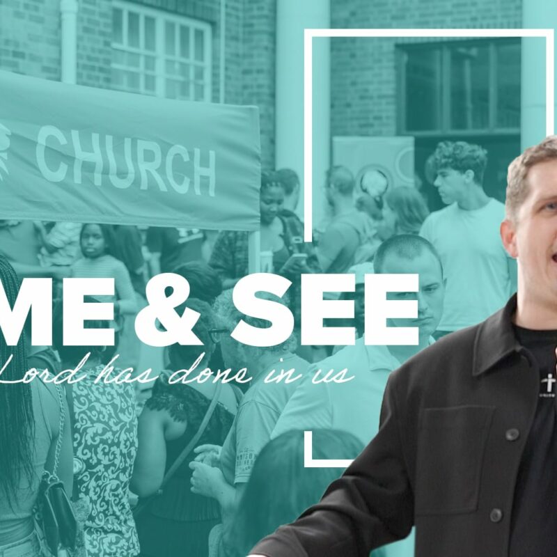 A photograph of a man with light brown hair and a black jacket, holding a microphone and speaking to a crowd. Behind him, a crowd of people are gathered under a tent with the words "FREEDOM CHURCH" on it. Text overlays the image, reading "COME & SEE what the Lord has done in us." The colours are a mixture of black, grey, white, and a blueish-green tint.