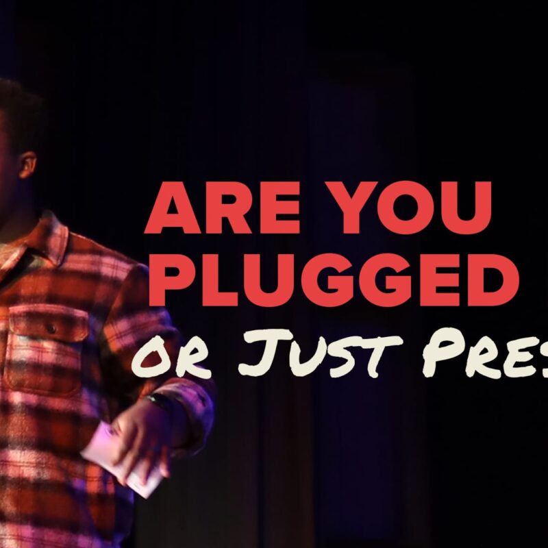 A photograph of a young man, Bradley Mubenga, wearing a plaid shirt and speaking passionately into a microphone on a dark stage. Large red and white text overlaid on the image reads: "ARE YOU PLUGGED IN OR JUST PRESENT?"