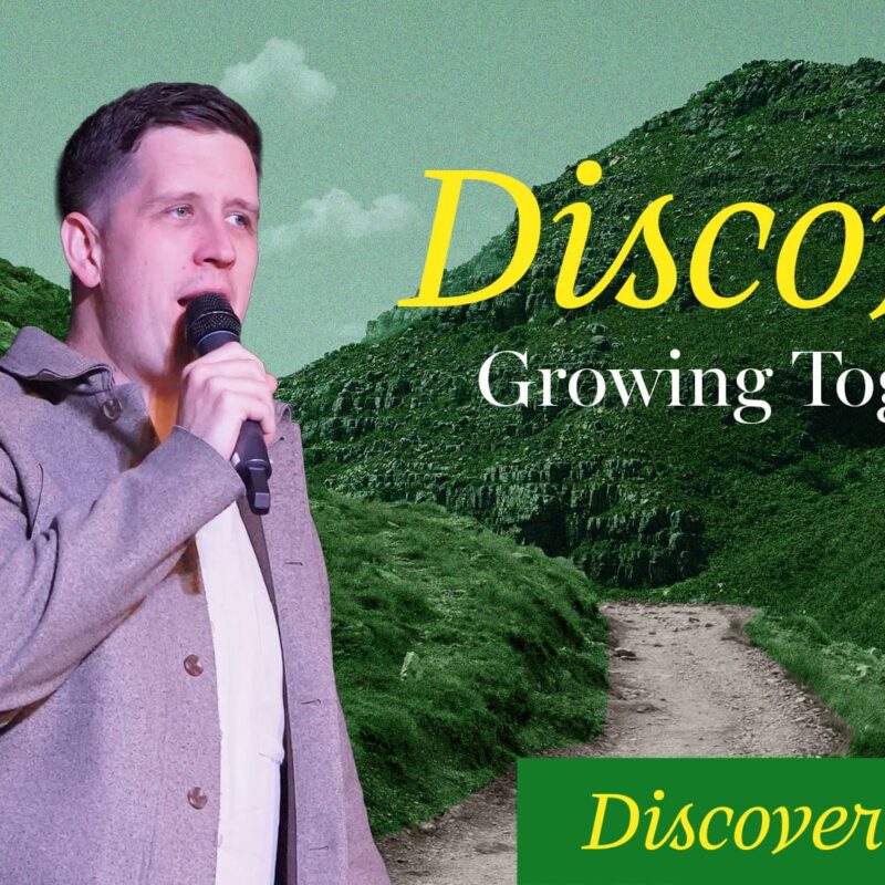 A man in a grey jacket holds a microphone, speaking, against a backdrop of green, rocky hills and a path leading up. The word "Discover" is written in large yellow script at the top. Below it, in white, are the words "Growing Together," and in a green box, "Discover Purpose."