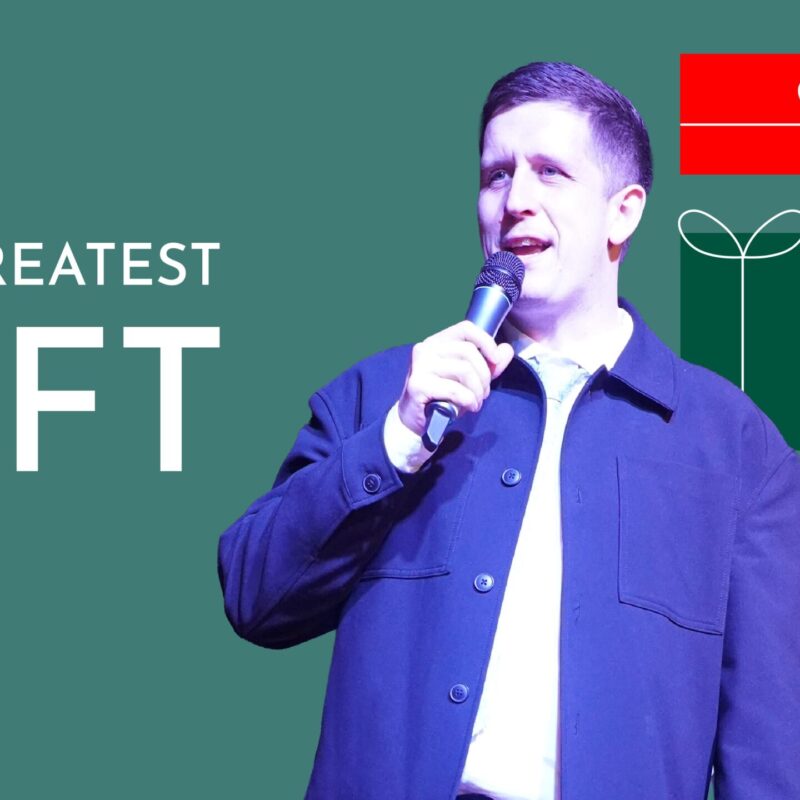 Pastor Josh Cooke speaking into a microphone on a stage. The graphic has a green background and is titled "THE GREATEST GIFT". To the side is a stack of stylized red, green, blue, and pink Christmas presents.