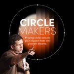 A man in a brown t-shirt stands on the left, pointing directly toward the viewer with his right hand while holding a small, open book in his left. He is positioned against a solid black background. To his right, a glowing white graphic features a "prayer circle" design. Inside the circle, the bold white text reads: "CIRCLE MAKERS: Praying circles around your biggest fears and greatest dreams." A bright lens flare effect glows at the top left of the circle.