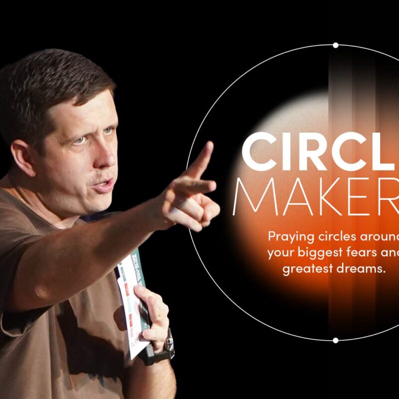 A man in a brown t-shirt stands on the left, pointing directly toward the viewer with his right hand while holding a small, open book in his left. He is positioned against a solid black background. To his right, a glowing white graphic features a "prayer circle" design. Inside the circle, the bold white text reads: "CIRCLE MAKERS: Praying circles around your biggest fears and greatest dreams." A bright lens flare effect glows at the top left of the circle.