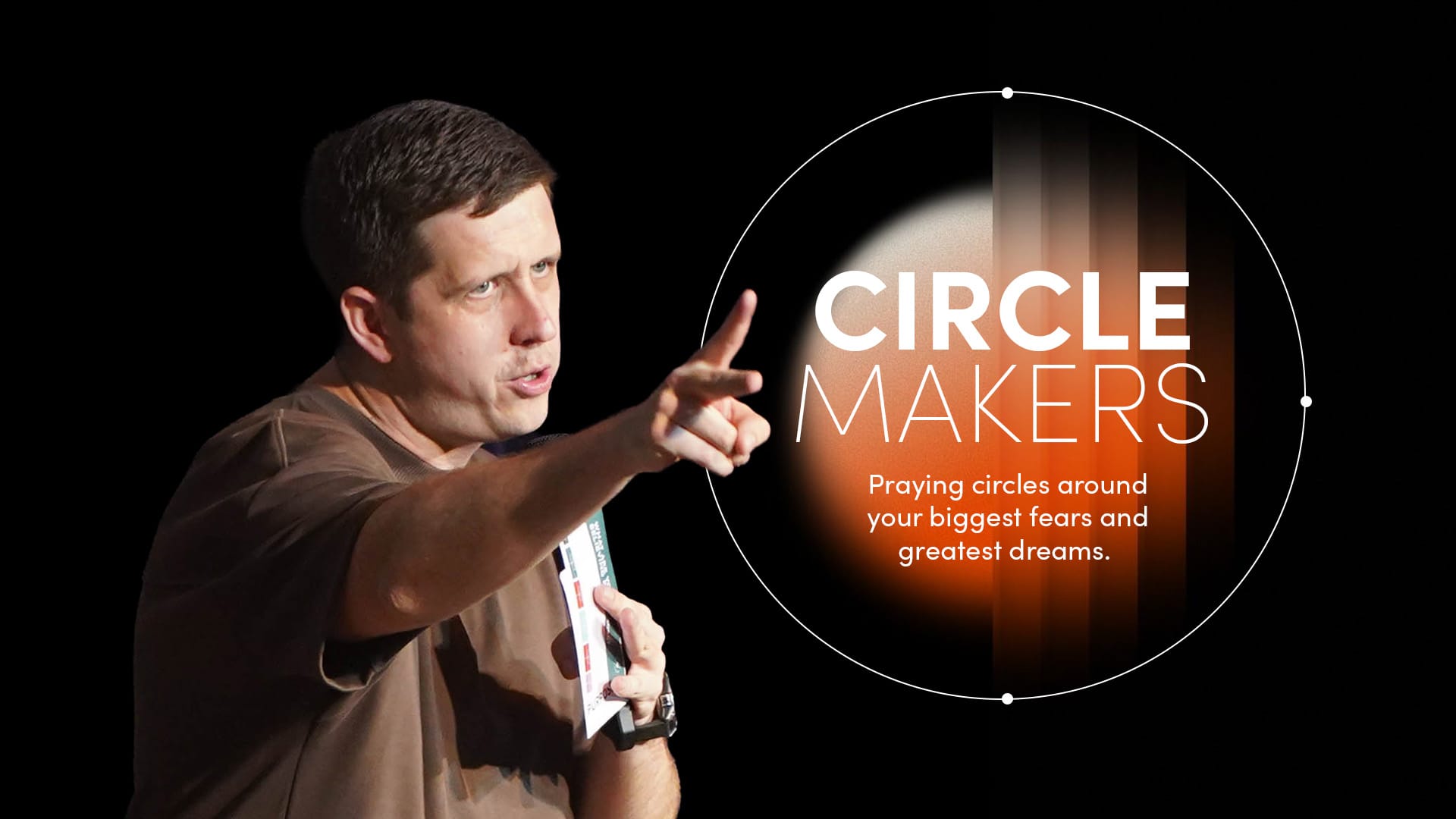 A man in a brown t-shirt stands on the left, pointing directly toward the viewer with his right hand while holding a small, open book in his left. He is positioned against a solid black background. To his right, a glowing white graphic features a "prayer circle" design. Inside the circle, the bold white text reads: "CIRCLE MAKERS: Praying circles around your biggest fears and greatest dreams." A bright lens flare effect glows at the top left of the circle.