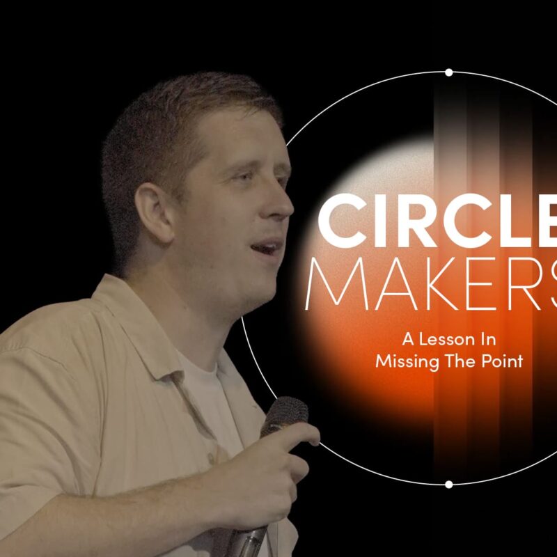 A medium shot of a man, identified as Pastor Josh Cooke, standing on a stage and speaking into a handheld microphone. He is wearing a light-coloured, short-sleeved button-down shirt over a white t-shirt and is holding a book, likely a Bible, in his other hand. To his right, a large graphic overlay features a white circular border with the words "CIRCLE MAKERS" in bold, white sans-serif lettering. Below the title, the text reads, "Praying circles around your biggest fears and greatest dreams". The background is dark with a soft, orange-to-black vertical gradient glow on the right side.