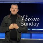 A promotional image for global vision Sunday. Pastor Gary stands on a stage lit in blue, wearing a black denim jacket over a black shirt with his arms meeting at his chest. Stylised text saying "Vision Sunday" appears to the right.