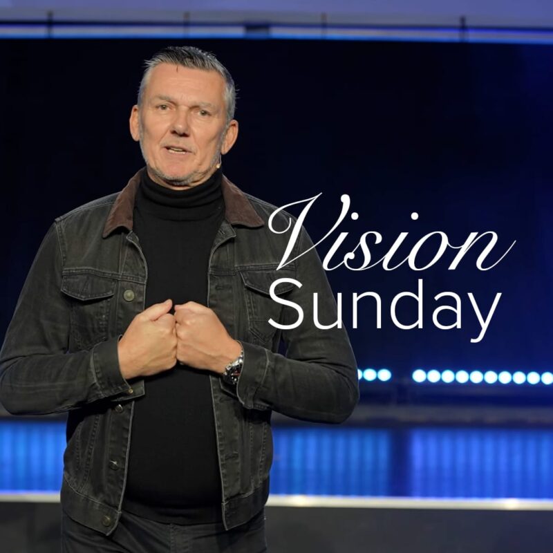 A promotional image for global vision Sunday. Pastor Gary stands on a stage lit in blue, wearing a black denim jacket over a black shirt with his arms meeting at his chest. Stylised text saying "Vision Sunday" appears to the right.